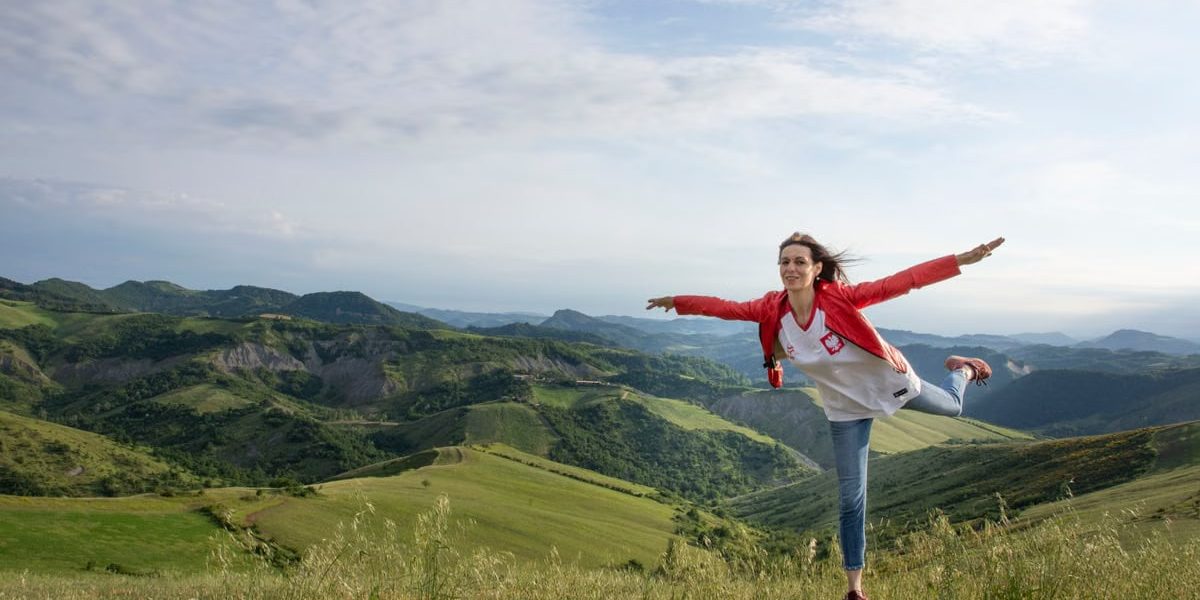 A woman joyfully balances on a rock amidst rolling green hills under a clear blue sky.