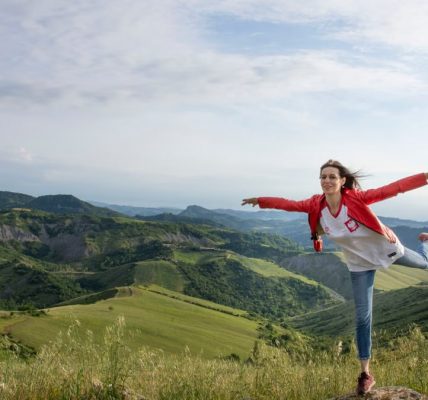 A woman joyfully balances on a rock amidst rolling green hills under a clear blue sky.
