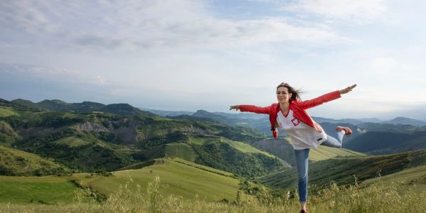 A woman joyfully balances on a rock amidst rolling green hills under a clear blue sky.